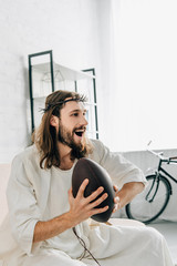 Canvas Print - side view of happy Jesus in crown of thorns holding ball and watching rugby on sofa at home