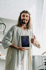 Canvas Print - smiling Jesus in crown of thorns doing thumb gesture and showing ipad tablet at home