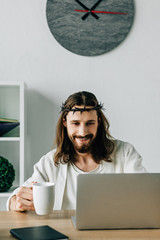 Wall Mural - cheerful Jesus in crown of thorns with coffee cup using laptop at table in modern office