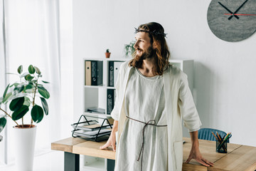 Wall Mural - selective focus of happy Jesus in crown of thorns and robe looking away near working table in modern office