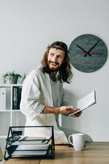 Wall Mural - angry Jesus in crown of thorns and robe sitting on table and pointing at textbook in modern office