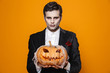 © Drobot Dean - Photo of scary dead man on halloween wearing classical suit and creepy makeup holding carved pumpkin, isolated over yellow background