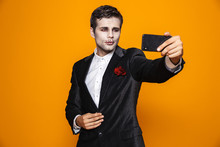 Photo Of Terrible Dead Man On Halloween Wearing Classical Suit And Creepy Makeup Taking Selfie On Mobile Phone, Isolated Over Yellow Background