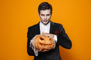 Photo of spooky dead man on halloween wearing classical suit and creepy makeup holding carved pumpkin, isolated over yellow background