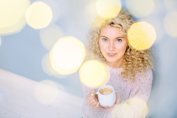 Attractive blond curly woman with a cup of coffee indoor over grey wall background. Photo taken through the garland light. Leisure, winter, holidays, people concept.