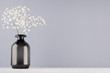 Soft light elegant home decor in minimalist style - black transparent vase with flowers branch on grey wall and white wood background.