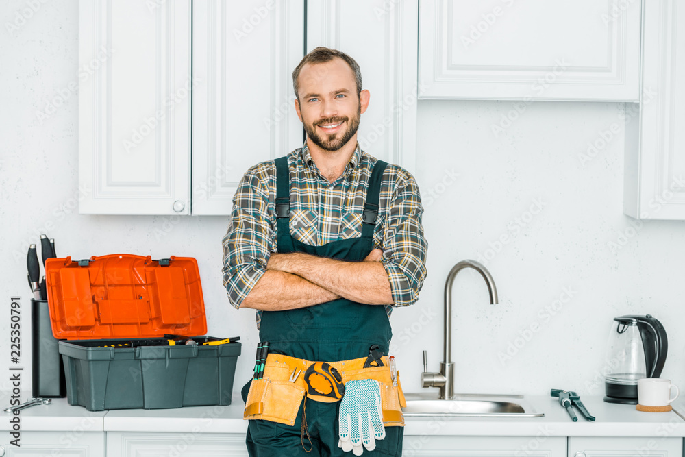 smiling handsome plumber standing with crossed arms and looking at camera in kitchen - obrazy, fototapety, plakaty 