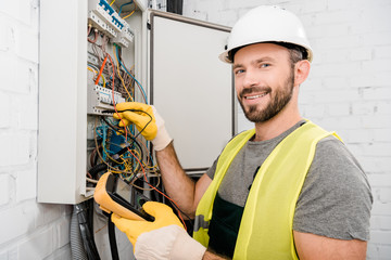 Canvas Print - smiling handsome electrician checking electrical box with multimetr in corridor and looking at camera