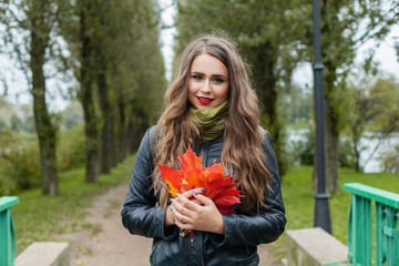 Happy young woman with long healthy hair, makeup and fall leaves in hand walking in autumn park