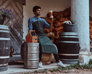 Wall Mural - Brewmaster sitting on a wooden barrel and holds a glass of craft beer, relaxes after work.