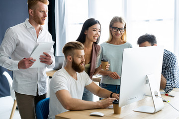 Wall Mural - Discussing successful project. Group of young cheerful business people working and communicating while sitting at the office desk together with colleagues sitting in the background.