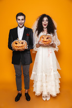 Full Length Photo Of Terrifying Zombie Couple Bridegroom And Bride Wearing Wedding Outfit And Halloween Makeup Holding Carved Pumpkin, Isolated Over Yellow Background