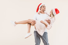 Happy Young Man In Santa Hat Carrying Beautiful Smiling Girl Isolated On Beige