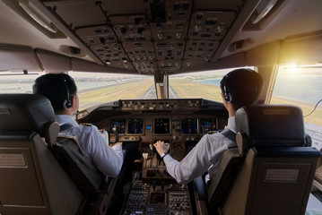 Two airliner pilots are flying the airplane towards the runway. Outside cockpit can see landing runway and environment with sun. Inside cockpit can see pilots and all flight instrument and equipment.