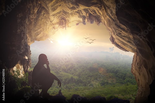 Fotomural Silhouette humble man sitting on a rock in cave looking at birds flying on autumn sunset background