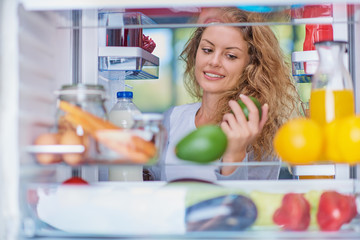 Wall Mural - Woman standing in front of opened fridge and taking avocado. Fridge full of groceries. Picture taken from the iside of fridge.