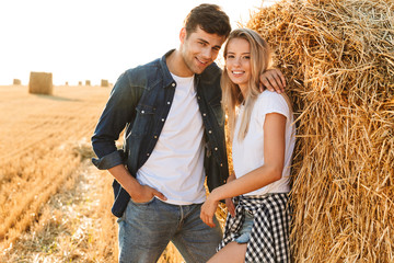 Image of smiling couple man and woman walking on golden field, and standing near big haystack