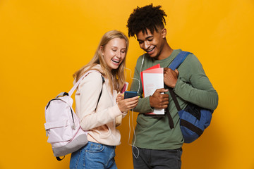 Photo of multiethnic students man and woman 16-18 wearing earphones using mobile phones and holding exercise books, isolated over yellow background