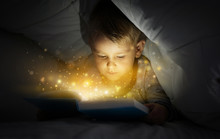 Cute Little Boy Reading Book In Bed Under Blanket