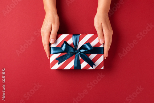 Female's hands holding striped gift box with blue ribbon on red background. C...