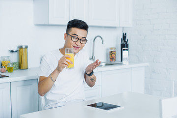 Wall Mural - happy young asian man in eyeglasses holding glass of juice and using digital tablet in kitchen