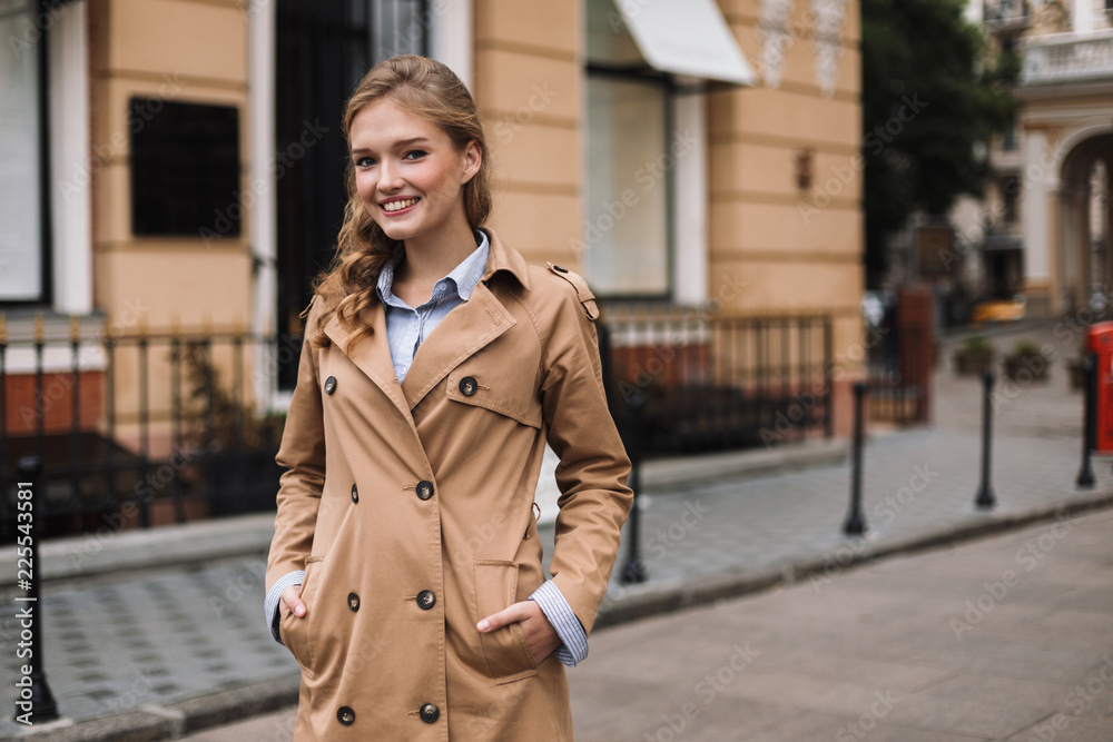 Young smiling woman in trench coat joyfully looking in camera while walking around cozy city street - obrazy, fototapety, plakaty 