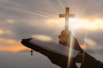 Silhouette of human hand holding the cross with bible, the background is the sunrise., Concept for Christian, Christianity, Catholic religion, divine, heavenly, celestial or god.