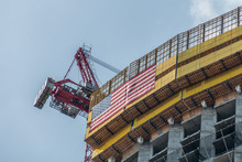 A Tower Crane Is Attached To Building Under Construction. This The Top Of The Building With A Large American Flag. A Glue Sky With A Small Cloud Is In The Background.