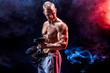 © zamuruev - Topless man exercising biceps with dumbbells posing in studio fuul of colored smoke