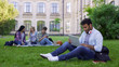 © motortion - Mixed race student using laptop, sitting on grass on campus, education online