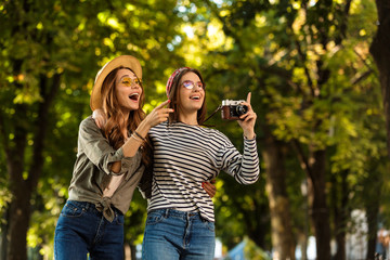 Sticker - Excited pretty young happy women friends walking outdoors with backpacks and camera pointing.