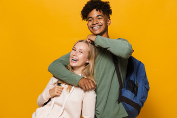 Wall Mural - Photo of teen people man and woman 16-18 wearing backpacks laughing and hugging together, isolated over yellow background