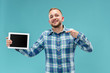 Studio picture of positive man isolated on blue background standing in casual clothes holding tablet and showing it blank screen with happy smile. He advising product or service.