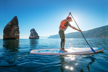 Young Man Having Fun Stand Up Paddling In The Sea. SUP. Guy Training In The Morning On Paddle Board Near The Rocks.