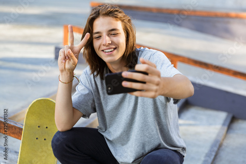 Fotografia Young skater guy sit in the park with skateboard take selfie by mobile phone with peace gesture