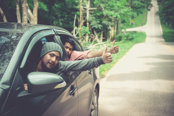 Men in car road trip waving out the window smiling. Two young Asian friend leaning out of car window.