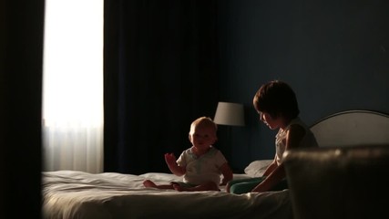 Wall Mural - Two sibling children, baby boy and his older brother, sitting on bed in sunny bedroom, playing together, back light