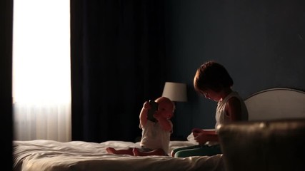 Canvas Print - Two sibling children, baby boy and his older brother, sitting on bed in sunny bedroom, playing together, back light