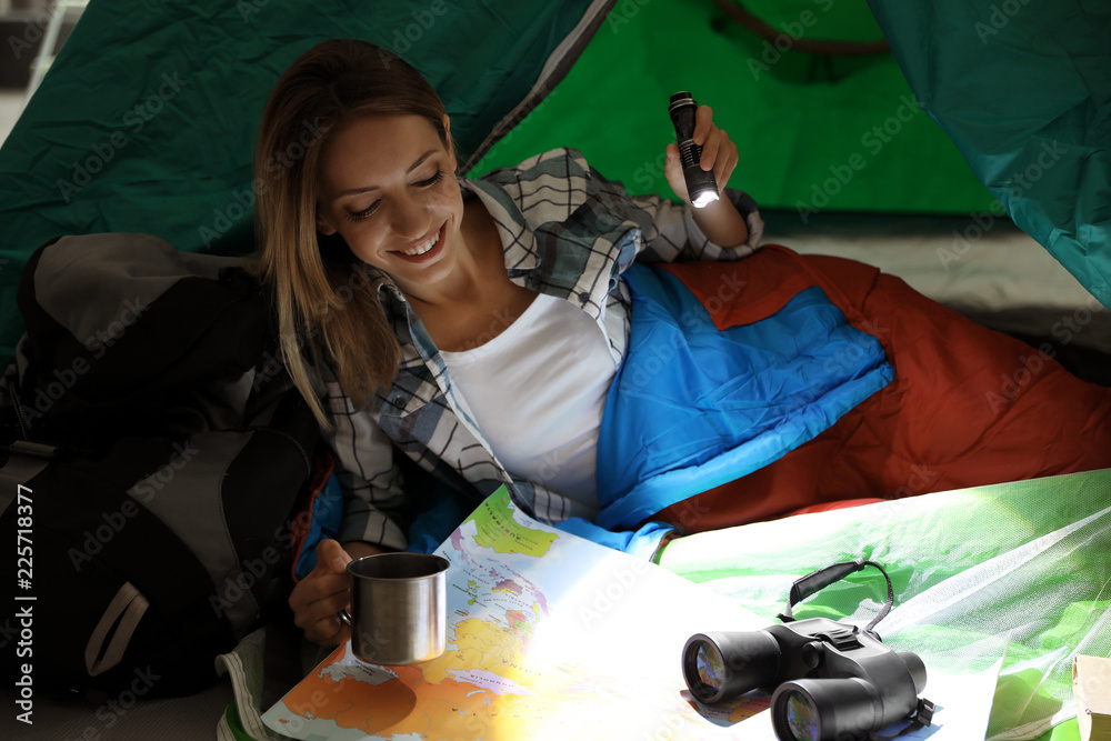 Young woman in sleeping bag with map and flashlight inside of tent - obrazy, fototapety, plakaty 