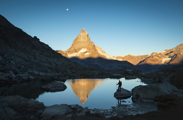 Fototapete - Hiker on a rock in the Riffelsee watching the first sunlight  on the Matterhorn.
