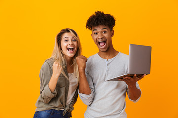 Poster - Happy young loving couple standing isolated over yellow background using laptop computer make winner gesture.