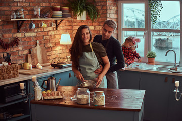 Wall Mural - Handsome male hug his wife while their little daughter sitting on window sill in loft style kitchen at morning.