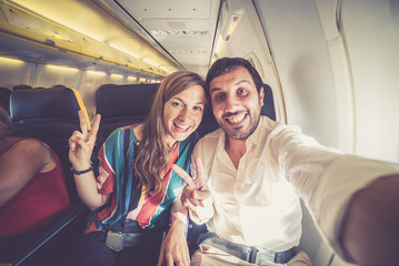 Wall Mural - Young handsome couple taking a selfie on the airplane during flight around the world. They are a man and a woman, smiling and looking at camera. Travel, happiness and lifestyle concepts.