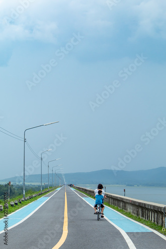 Fotografia, Obraz Mother and son ride this road on the reservoir.