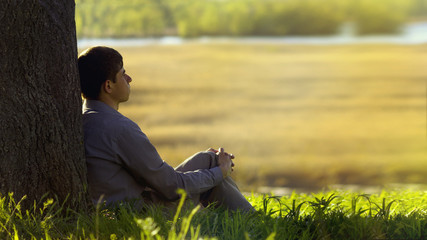 young man sitting with his back to the tree and meditating about life, the concept of religion and rest