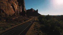 Drone Camera Follows Car Driving Along Small Desert Highway Road Near Breathtaking Steep Canyon Ridge On Sunset In USA.