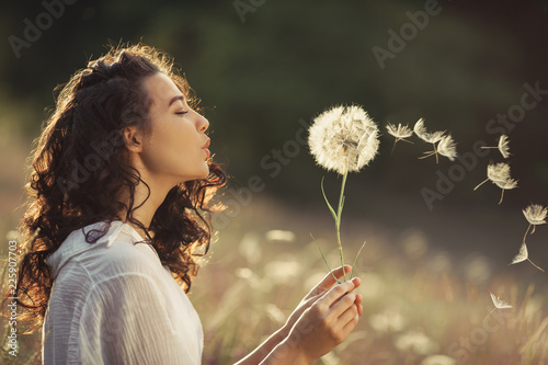 Leinwand Poster  Beautiful Young Woman sitting on the field in green grass and blowing dandelion