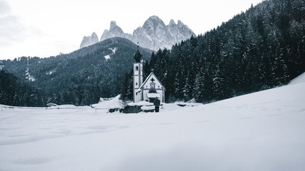 Church and houses in the village St. Magdalena in the Villnoesstal (Val di Funes) with the Geisler Dolomites mountains in the background and trees on the hills under a snow cover in winter, Italy.