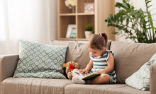 Childhood And People Concept - Little Girl Sitting On Sofa Reading Book And Toy Teddy Bear At Home
