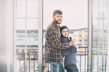 Young beautiful and happy couple man and woman at home on the balcony smiling and hugging on the background of city roofs